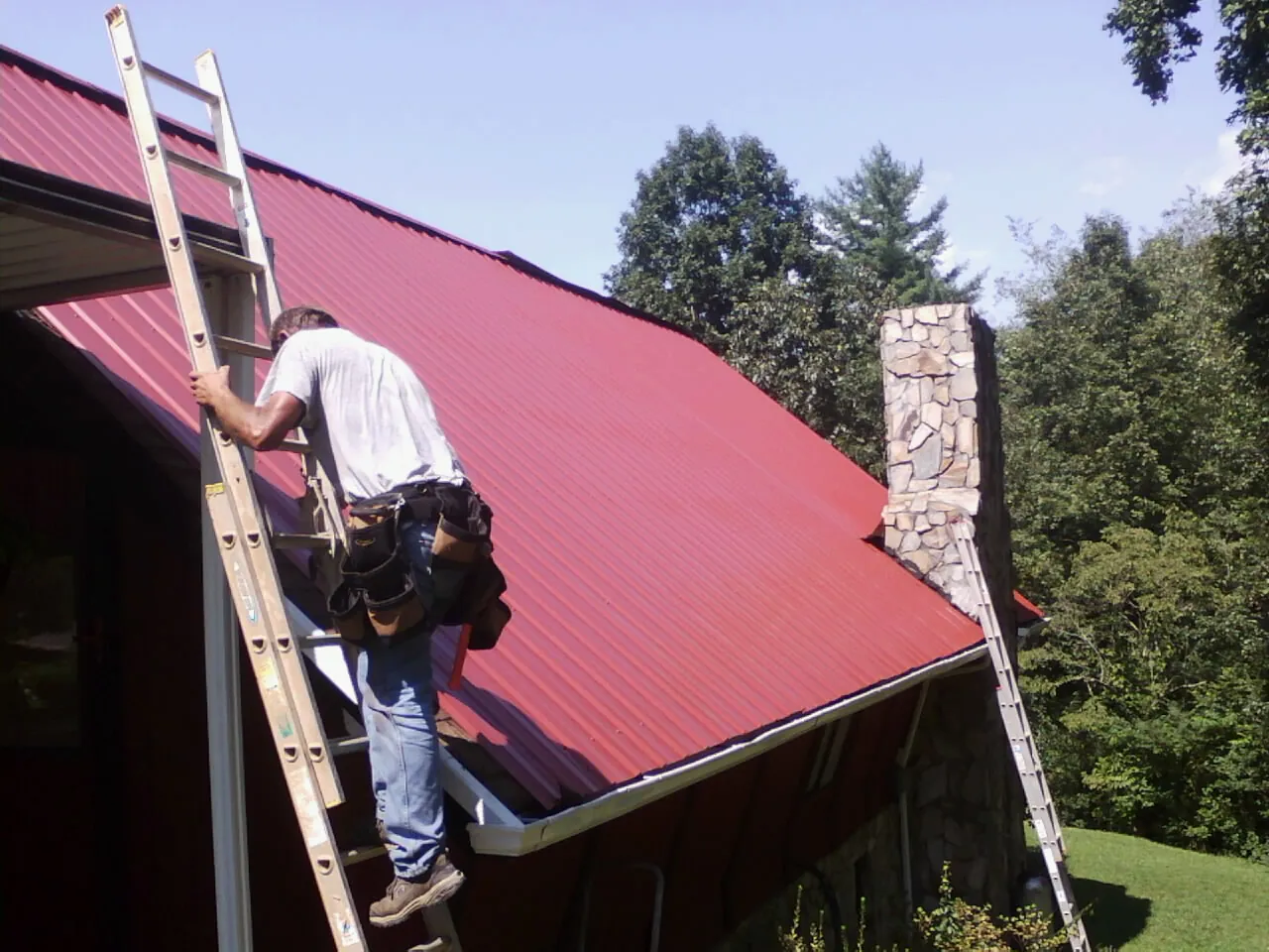 Roofer on a ladder beside a red metal roof and stone chimney — Soffit & Fascia Repair by Roofing Brothers in Barto